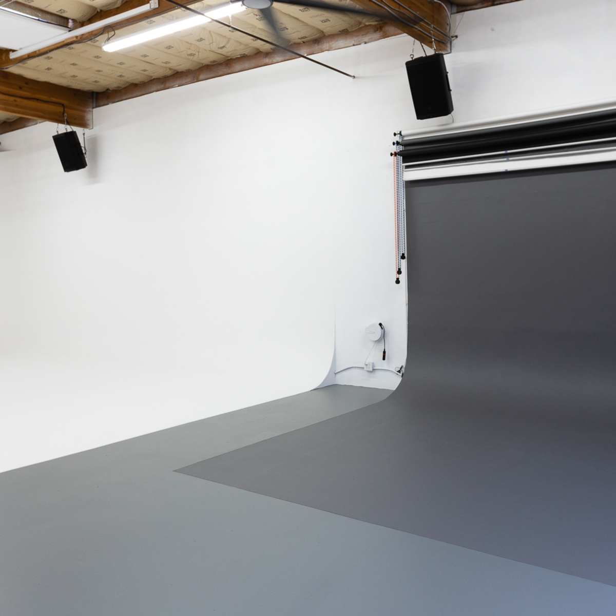 A minimalist photography studio with a white backdrop on one side and a gray backdrop on the other, featuring industrial ceiling beams and overhead lighting.