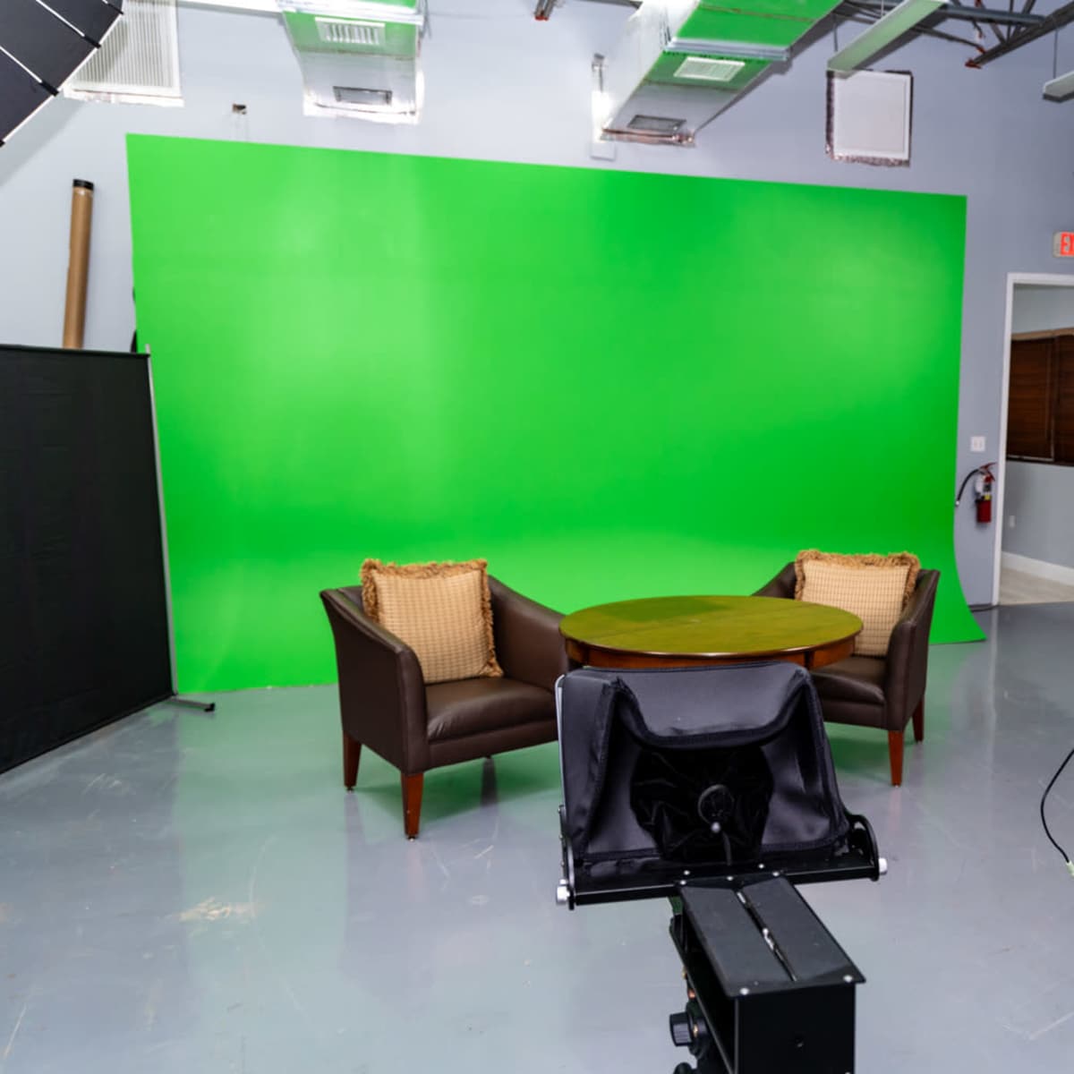 A video studio featuring two brown armchairs and a round table against a bright green background, with camera equipment positioned in the foreground.