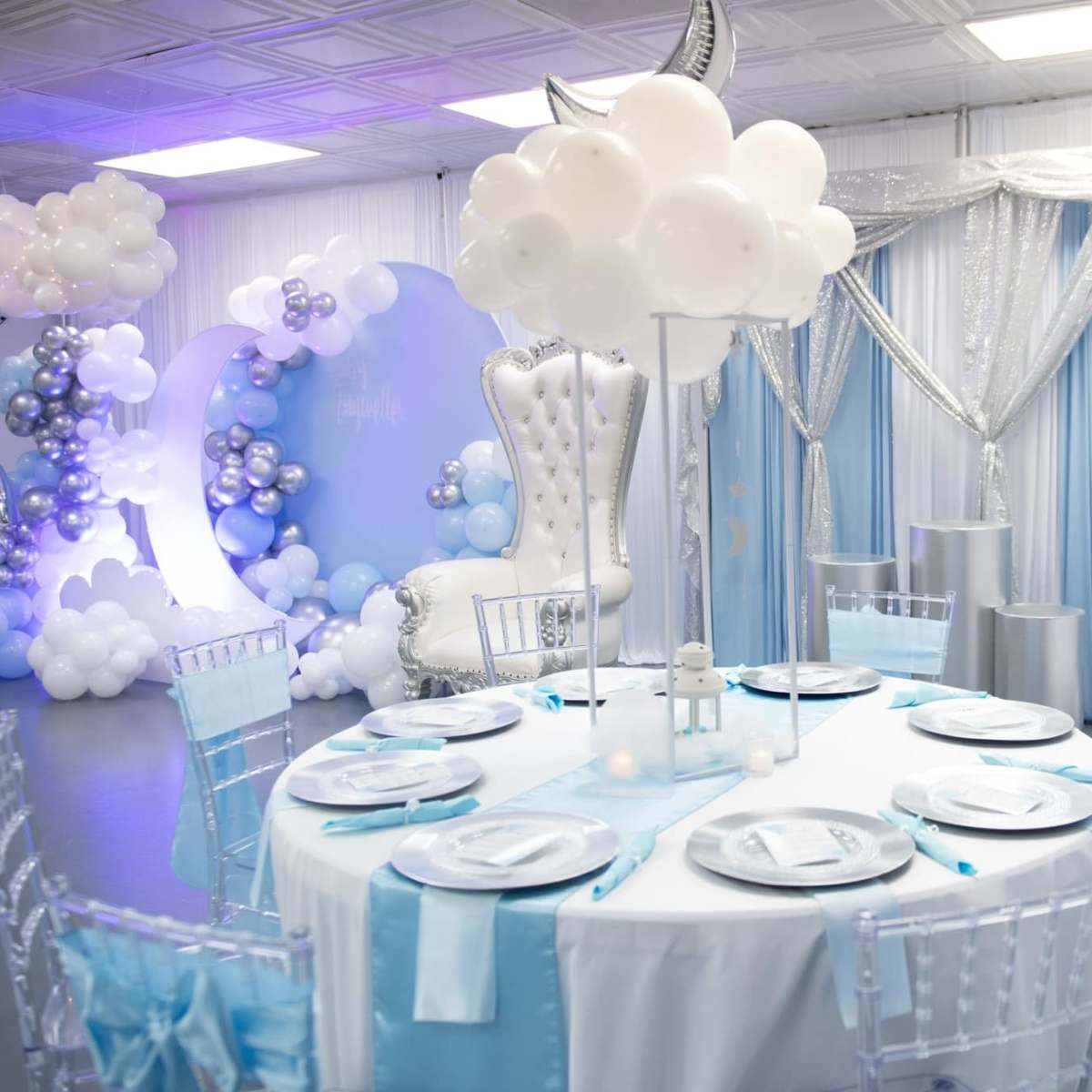 A decorated event space with a circular table set with light blue and white tableware, surrounded by elegant chairs, and a backdrop of balloon decorations and draped fabric.