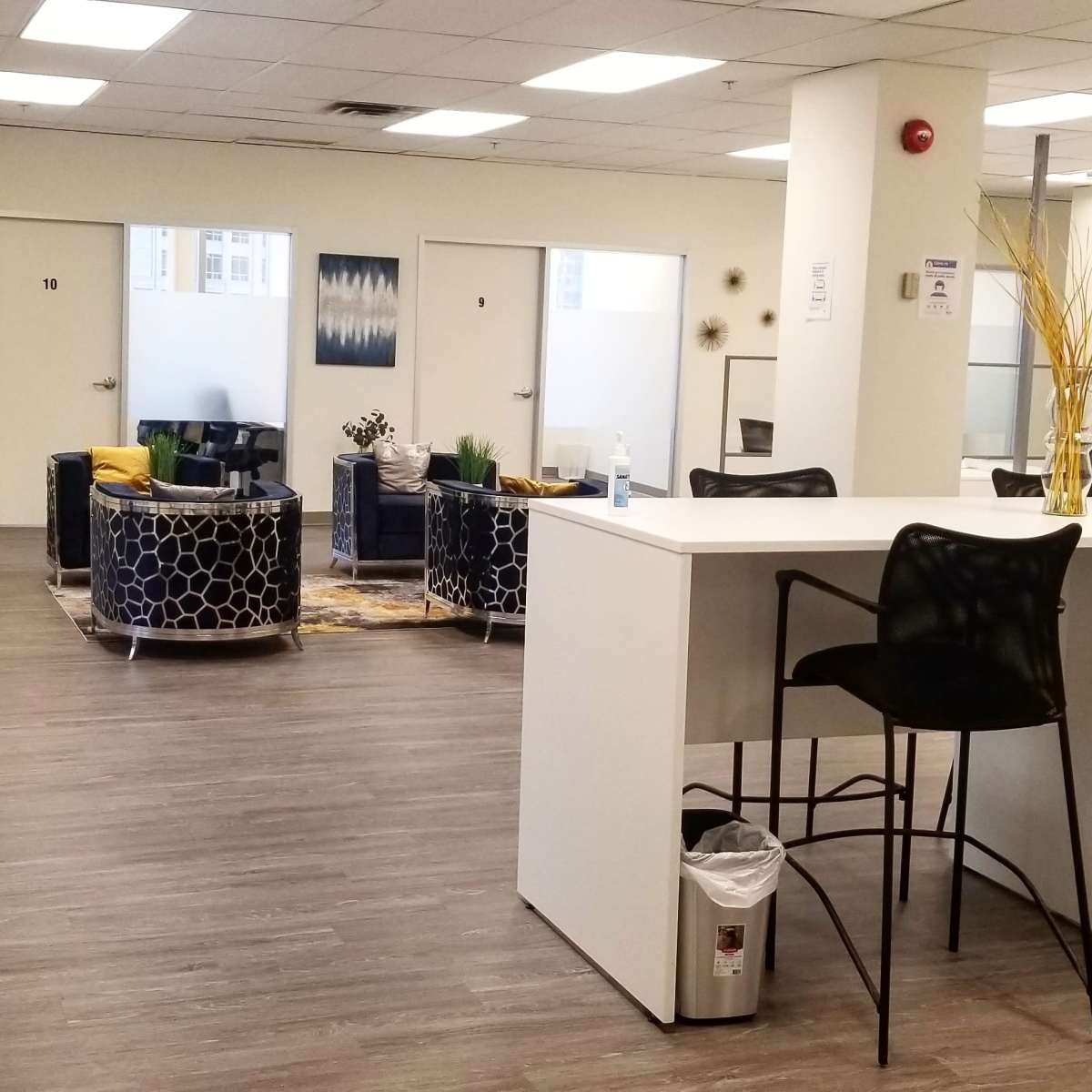 A modern office space with a kitchenette area and seating arrangements, featuring black and yellow chairs, a coffee table, and plants.