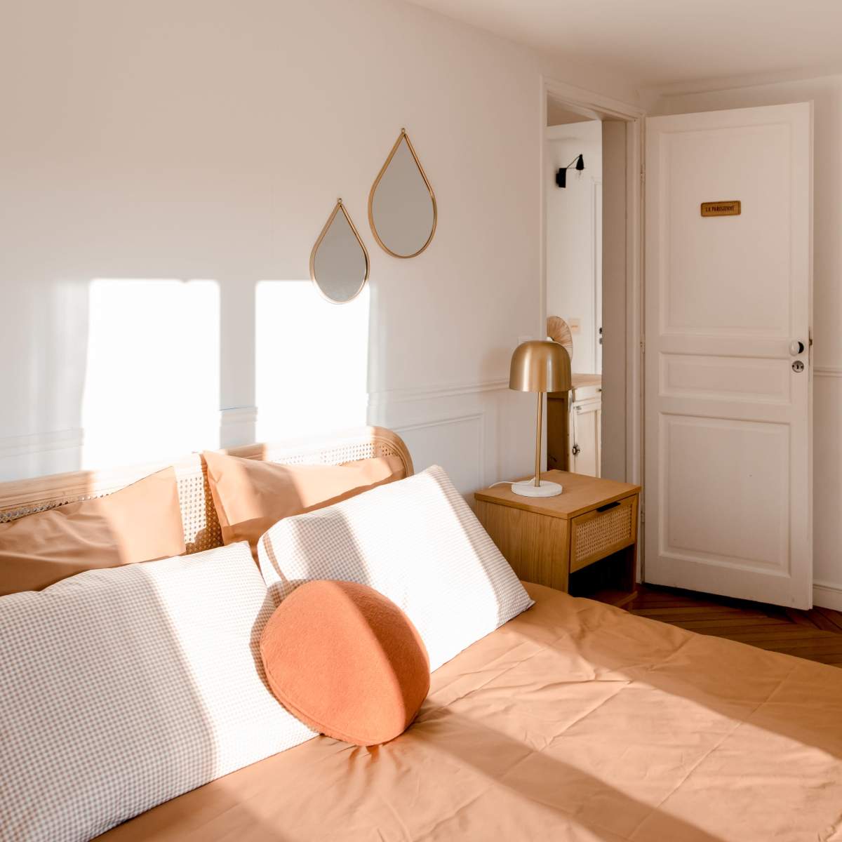 A softly lit bedroom featuring a bed with an orange pillow, a wooden bedside table, and two decorative mirrors on the wall.