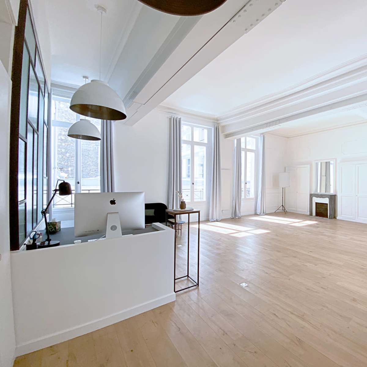 A bright, spacious room with light wooden flooring, large windows, and minimalist furniture, including a desk with a computer and a mirror.