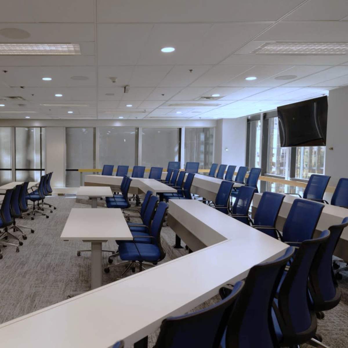 A modern conference room with a circular arrangement of white tables and blue chairs, equipped with large windows and two screens on the walls.