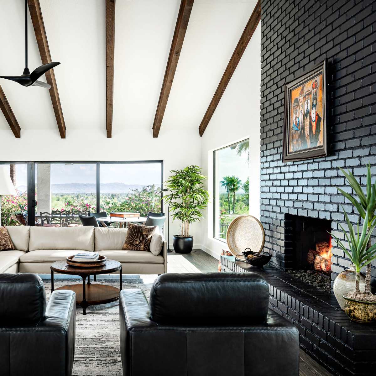 A modern living room with a high ceiling, featuring a sectional sofa, two black chairs, a stone fireplace, and large windows overlooking a garden.