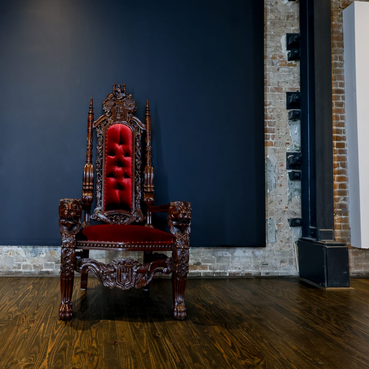 A dark wood throne with red upholstery is centered in front of a large black wall in an industrial-style room.