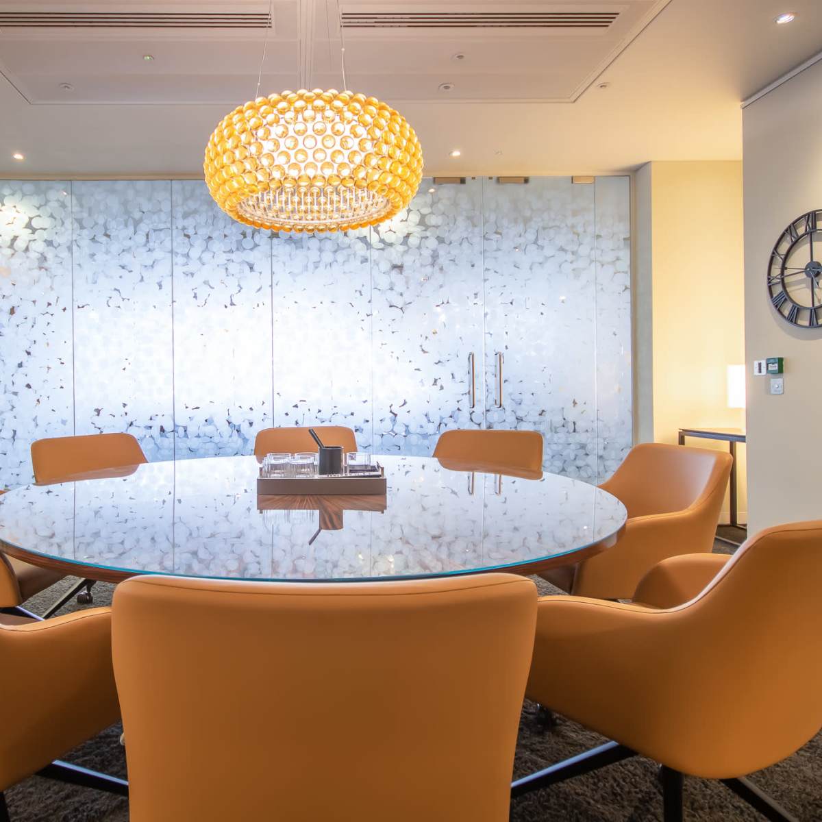 A round glass meeting table surrounded by orange chairs is set in a modern room with a decorative light fixture and a patterned glass wall.