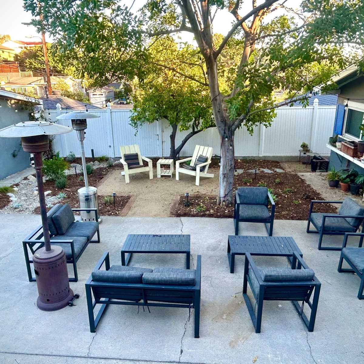 A backyard patio features multiple outdoor seating arrangements, a heater, and a tree centered in the garden area.