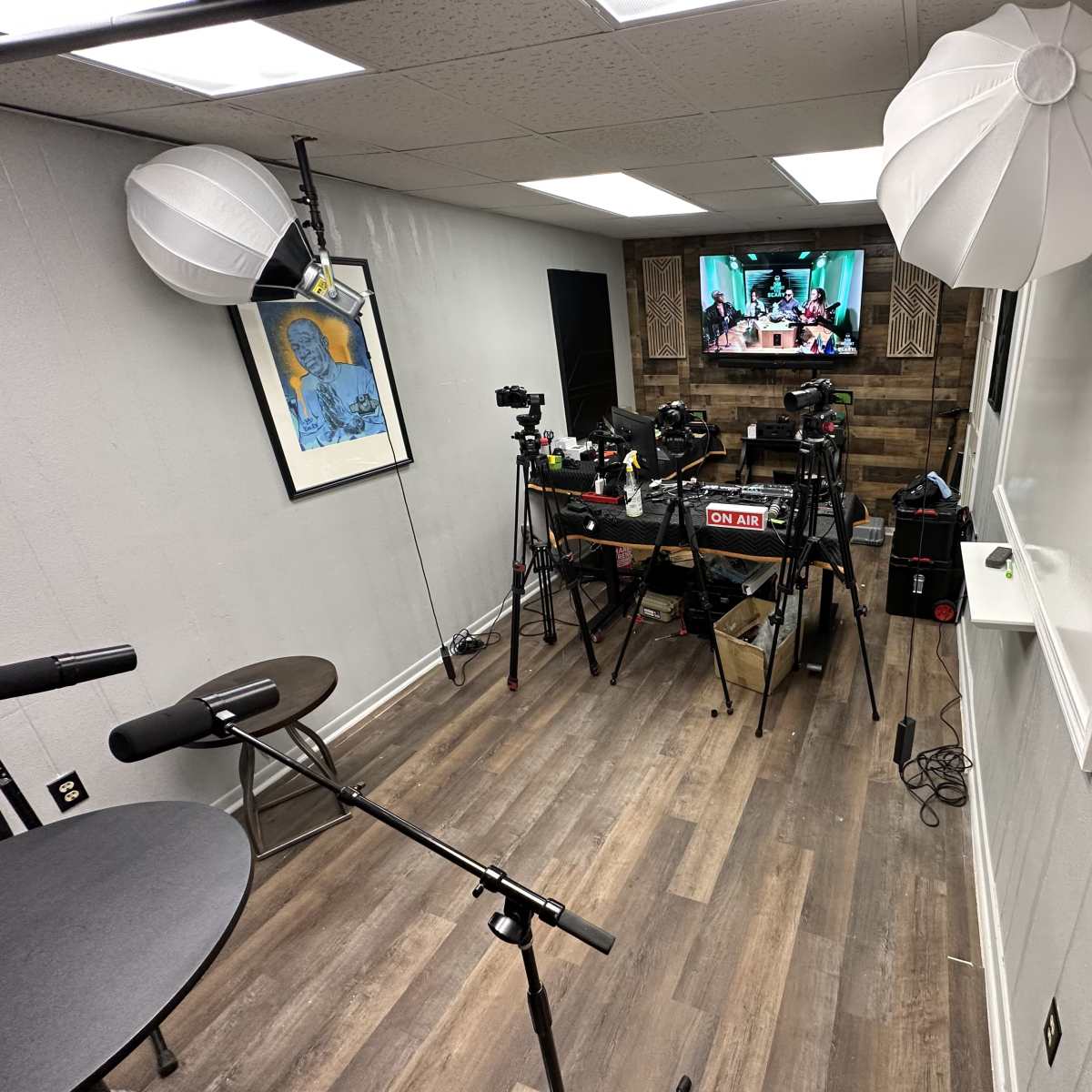 A small studio setup with multiple cameras, microphones, and a television screen displaying a group, all situated in a room with wooden panels and white walls.