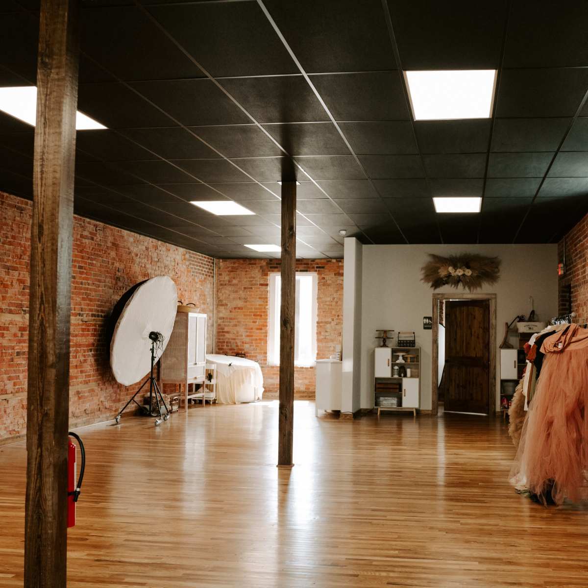An expansive studio space with exposed brick walls, wooden flooring, and creative props, including a large circular reflector and several costumes.
