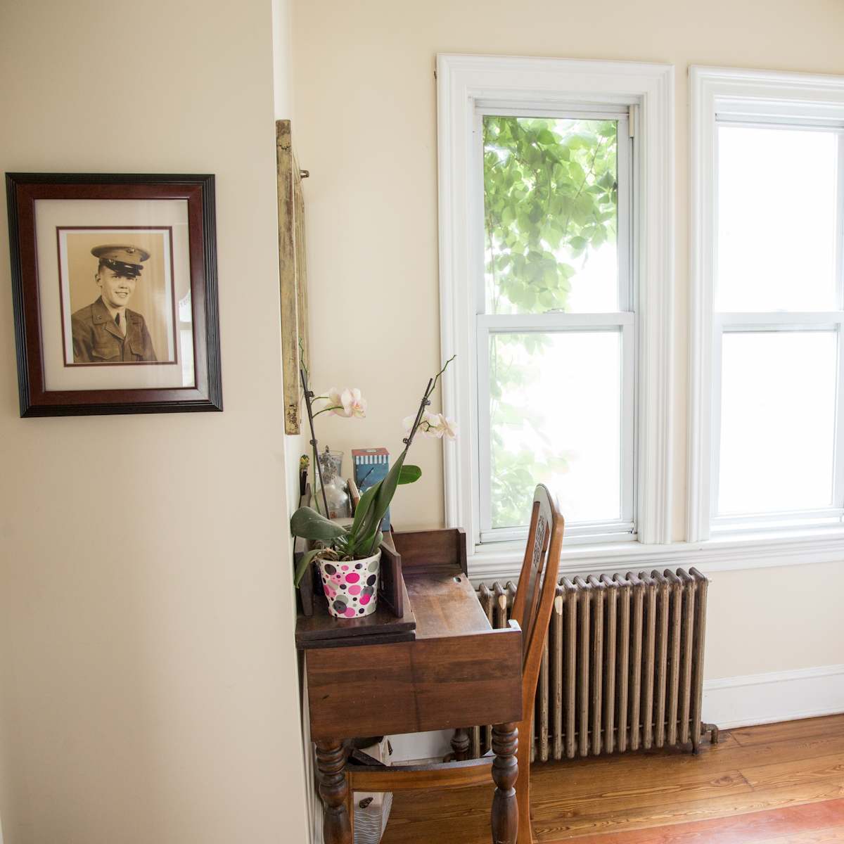 A small corner of a room featuring a wooden desk next to a radiator, framed by two windows and a framed portrait on the wall.