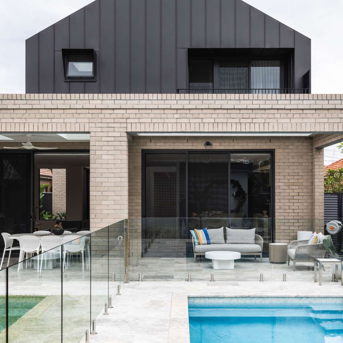 A modern house with a dark, sloped roof and large glass doors overlooks a swimming pool surrounded by a glass railing.