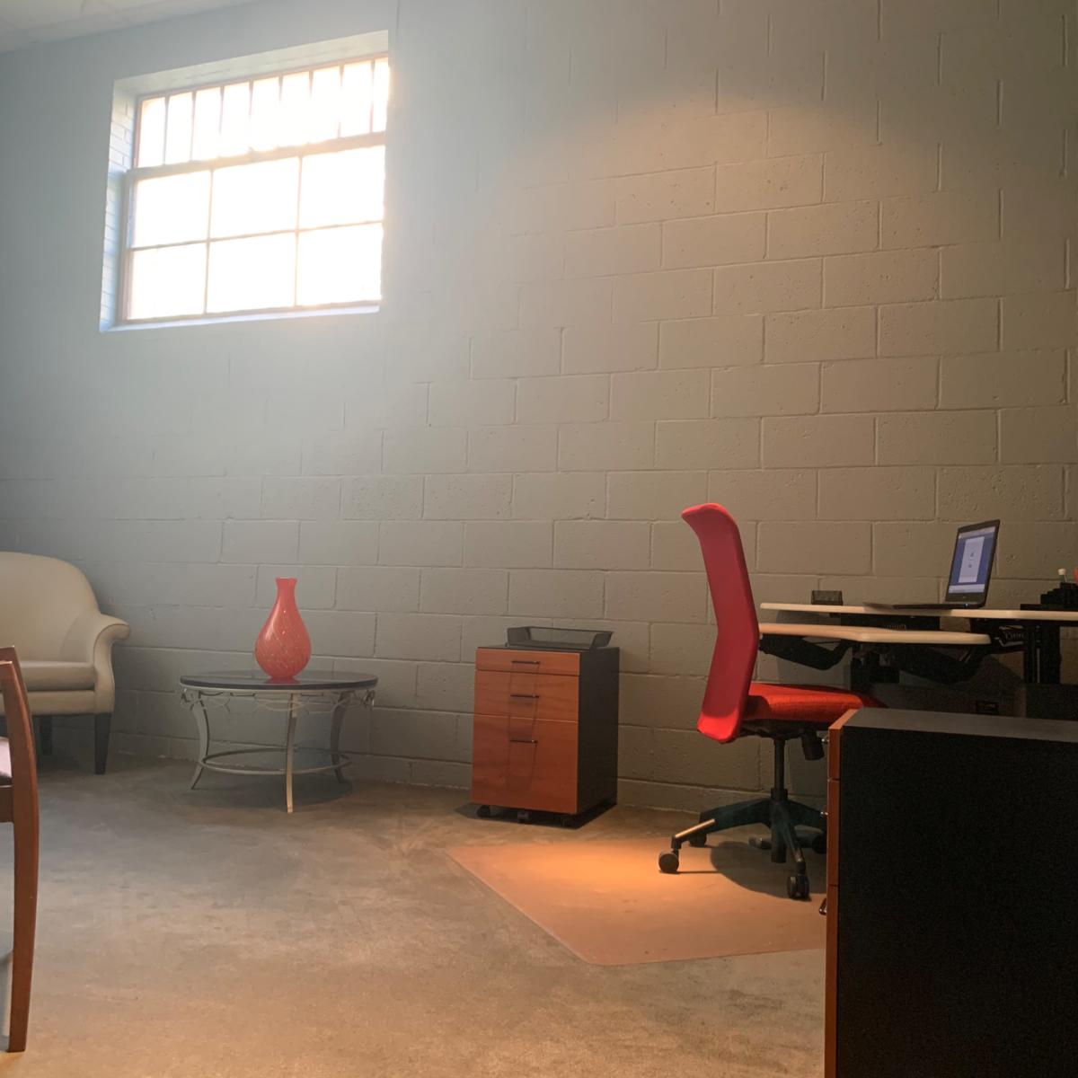 A sparsely furnished office with a desk, a red chair, a small table, an armchair, and a window letting in natural light.