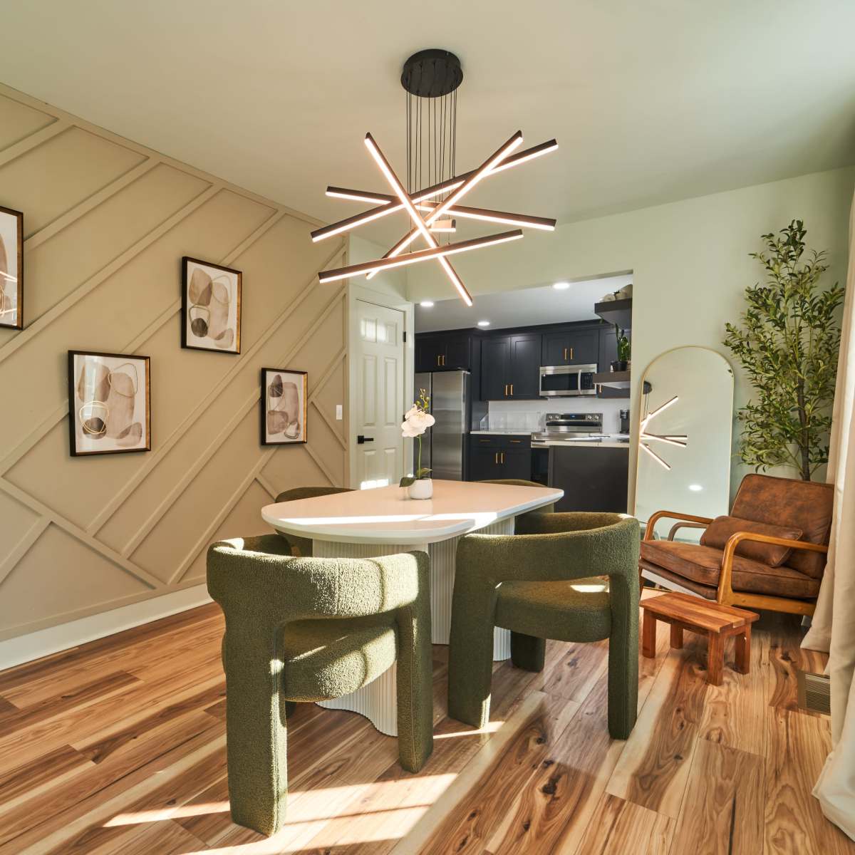 A modern dining area featuring a round table surrounded by green chairs, a unique light fixture, and decorative wall art, with a kitchen visible in the background.