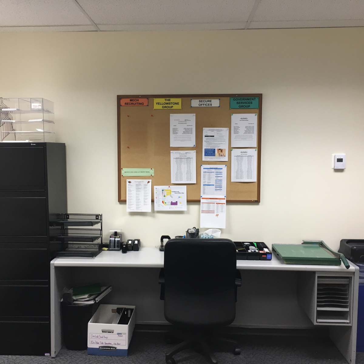 A small office with a desk, an office chair, a filing cabinet, and a bulletin board displaying various documents and notices.