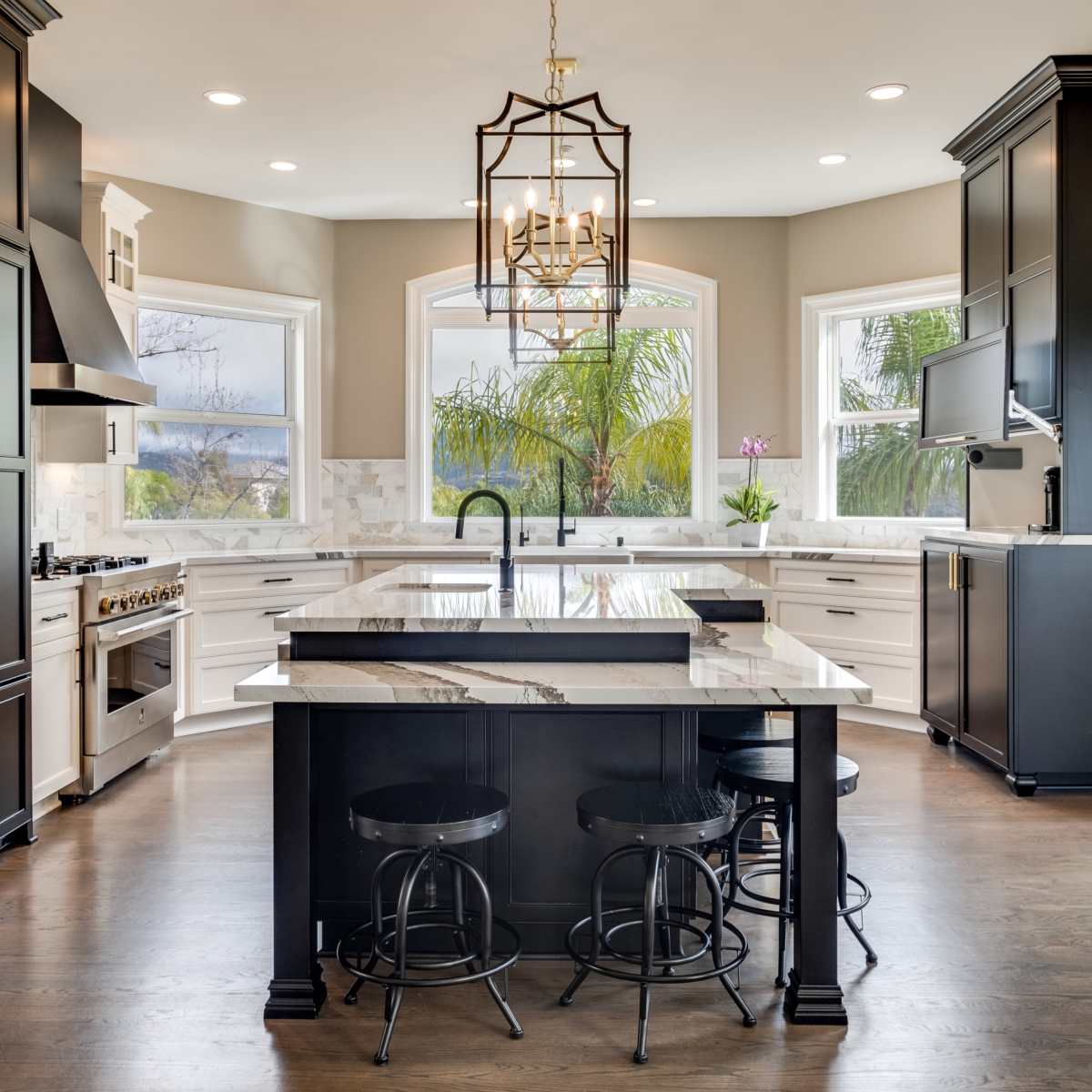 A modern kitchen features a central island with bar stools, white cabinetry, and large windows overlooking greenery.