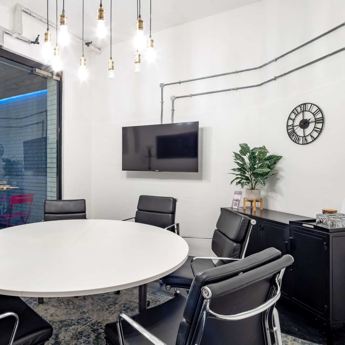A modern conference room features a round white table surrounded by black leather chairs, a flat-screen TV on the wall, and industrial-style lighting above.