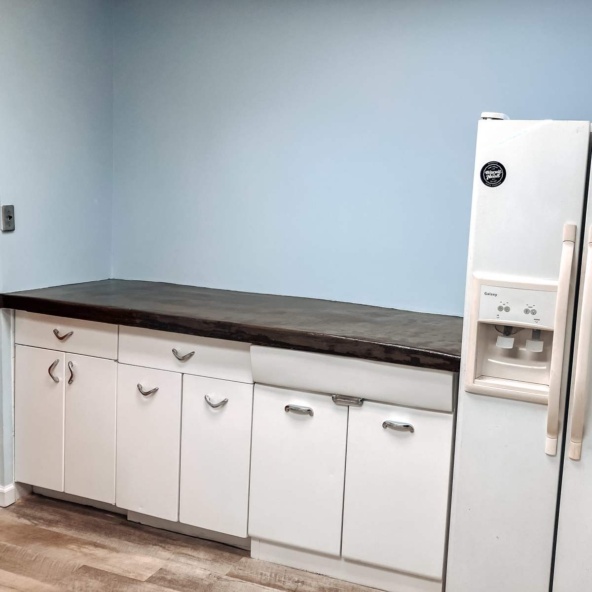 A kitchen space features a blue wall, a countertop, and white cabinetry with a side-by-side refrigerator.