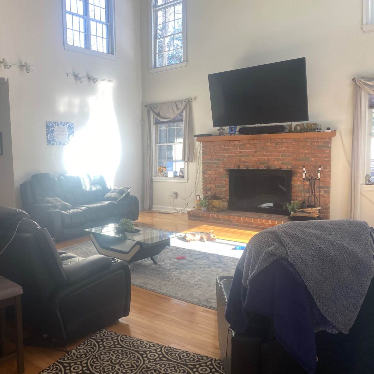 A spacious living room features leather sofas, a coffee table, and a brick fireplace with a large television mounted above it.