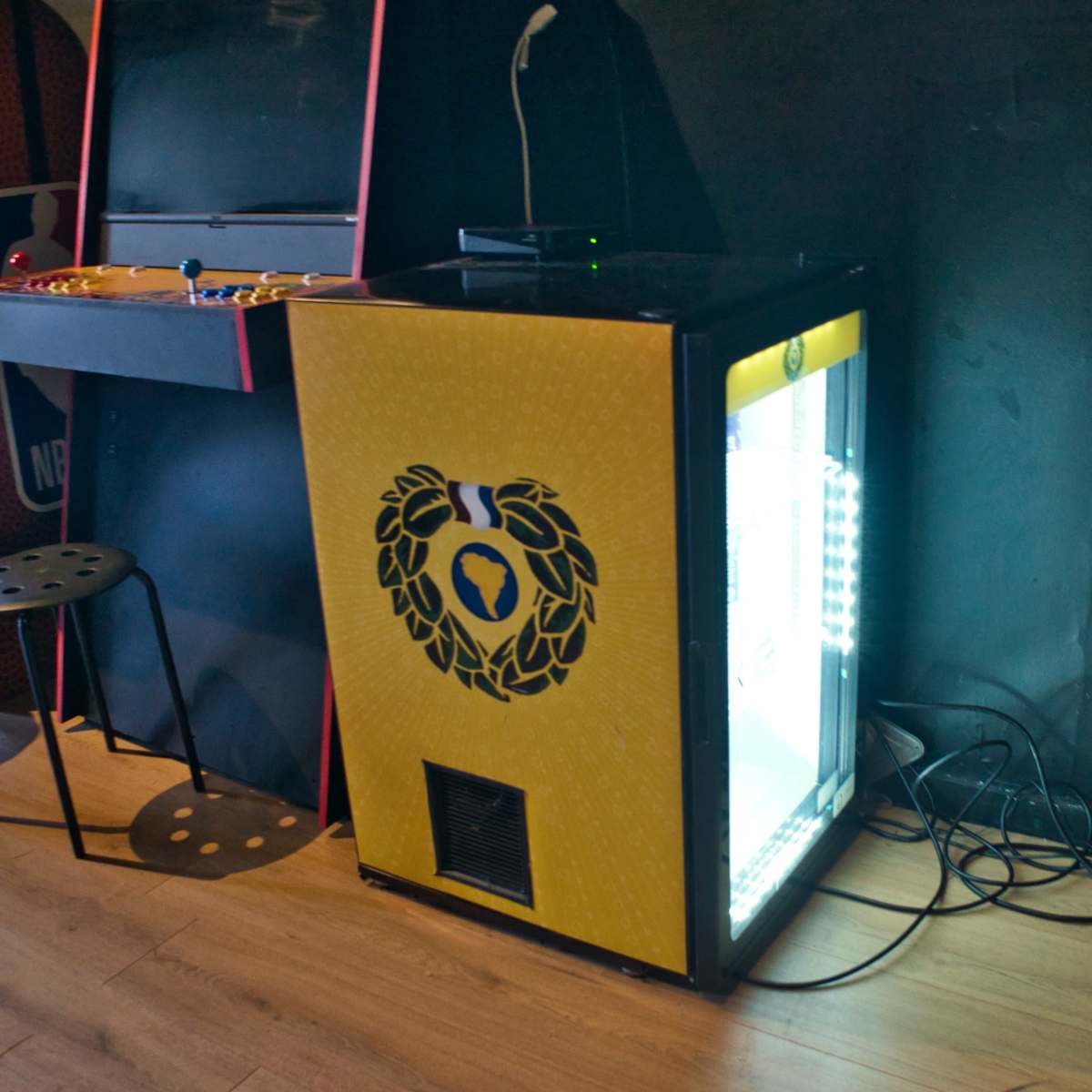 A vending machine with a yellow exterior and a glass front, illuminated from within, positioned next to arcade cabinets in a casual setting.
