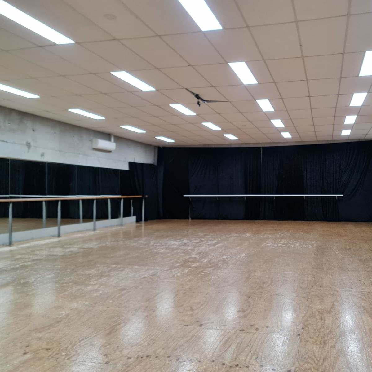 A spacious dance studio with a wooden floor, mirrors on one wall, and black curtains along another.