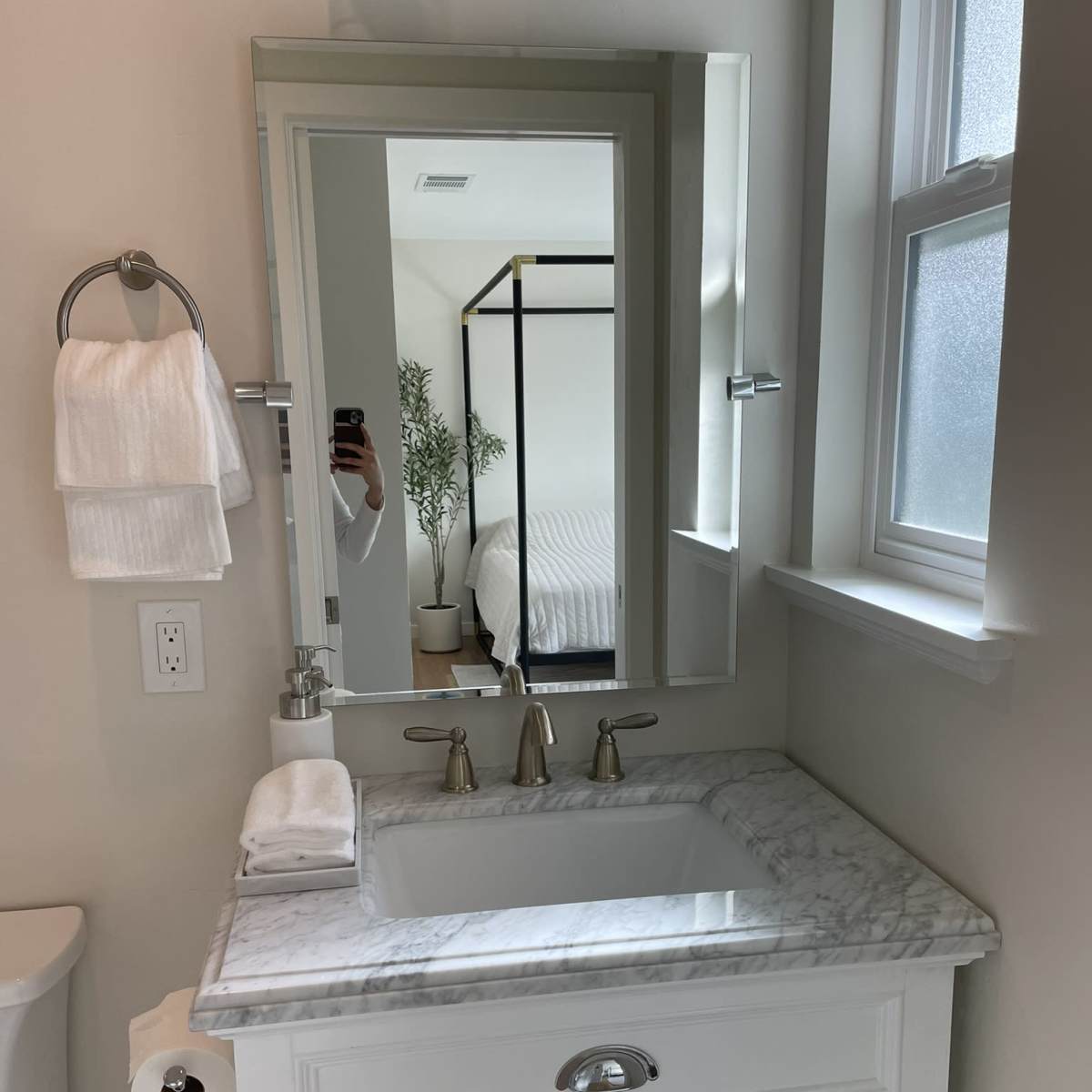 A white bathroom features a marble countertop with a sink, a large mirror, and a towel rack, with a glimpse of a bedroom visible in the background.