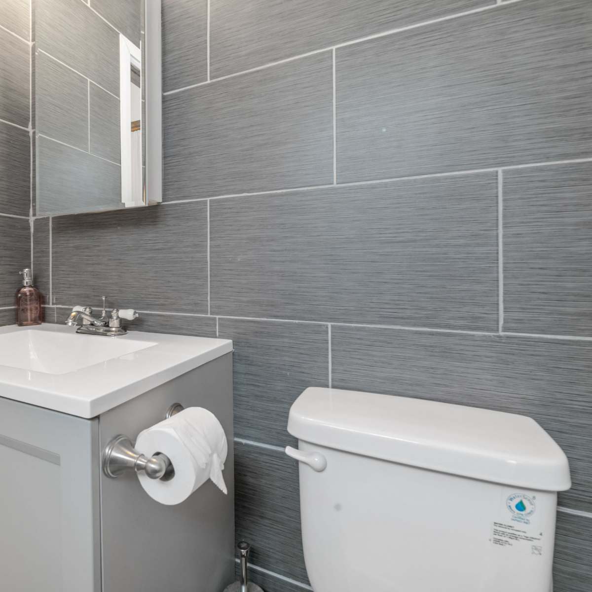 A modern bathroom featuring a gray tiled wall, a white sink cabinet with a mirror above, and a white toilet beside it.