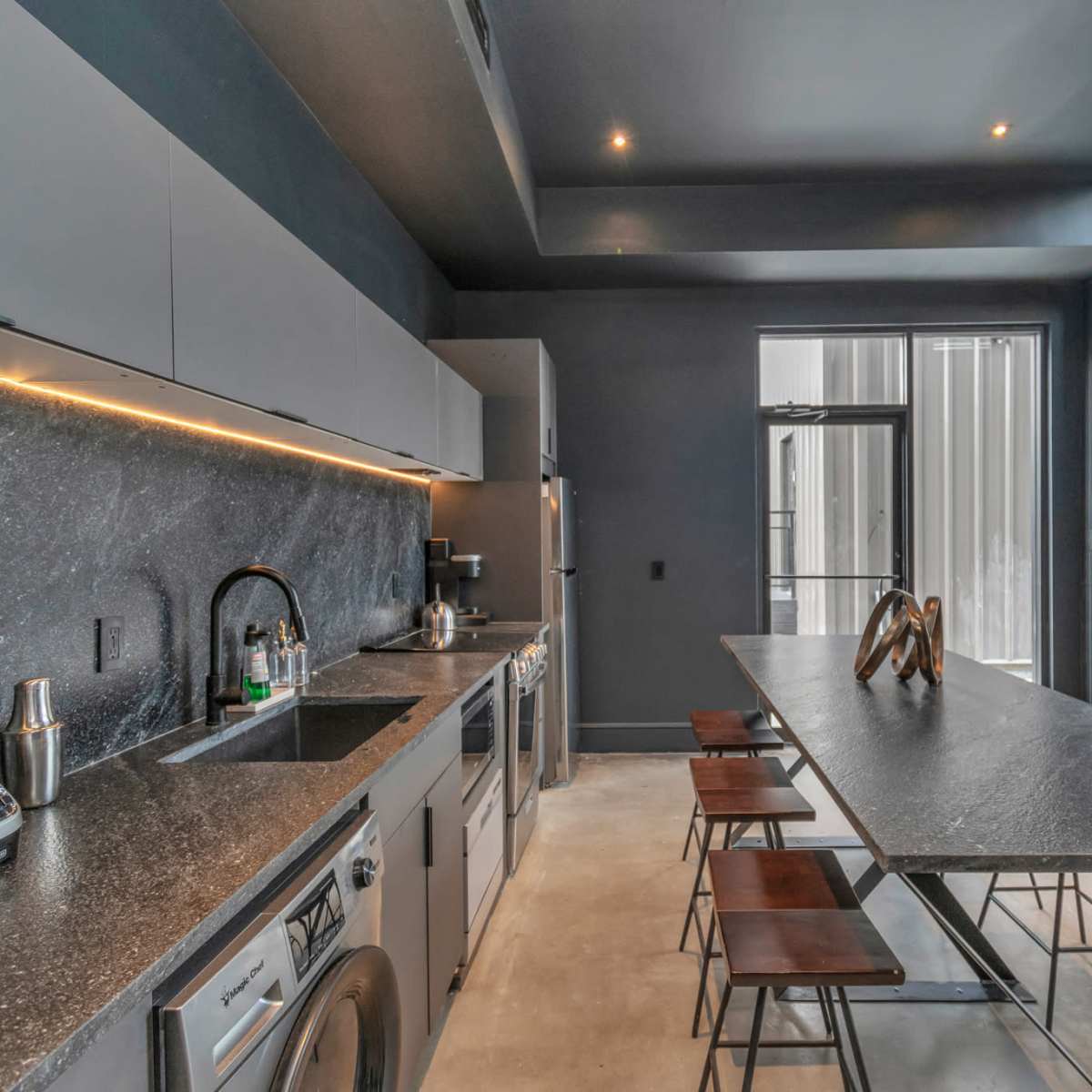 A modern kitchen features sleek gray cabinetry, a dark countertop, stainless steel appliances, and a minimalist dining table.