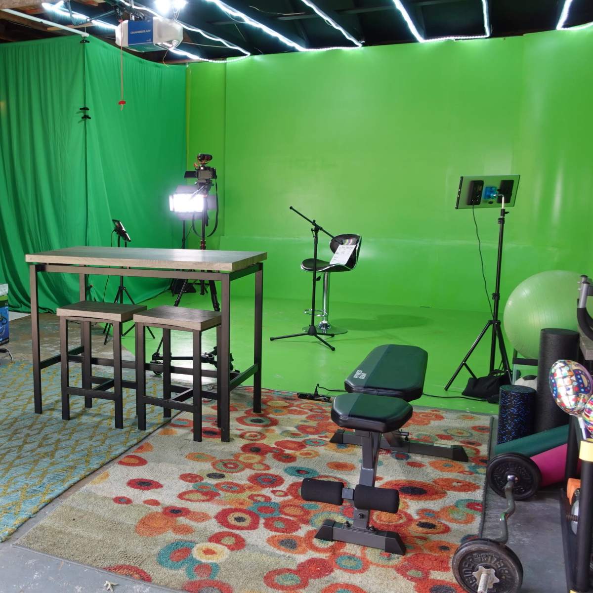 A workspace with a green screen setup, a desk with two stools, a chair, and various equipment arranged in a garage.