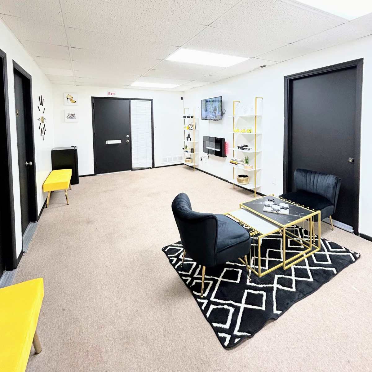 A modern waiting area featuring black chairs, a gold coffee table, a television on the wall, and decorative shelves, all set against a light-colored carpeted floor.