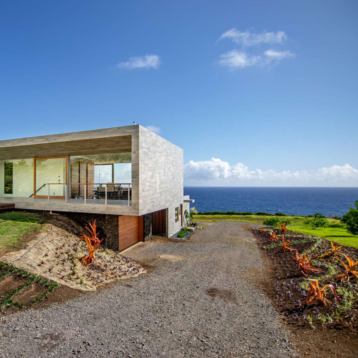 A modern concrete house with large glass windows is situated on a grassy hill overlooking the ocean.