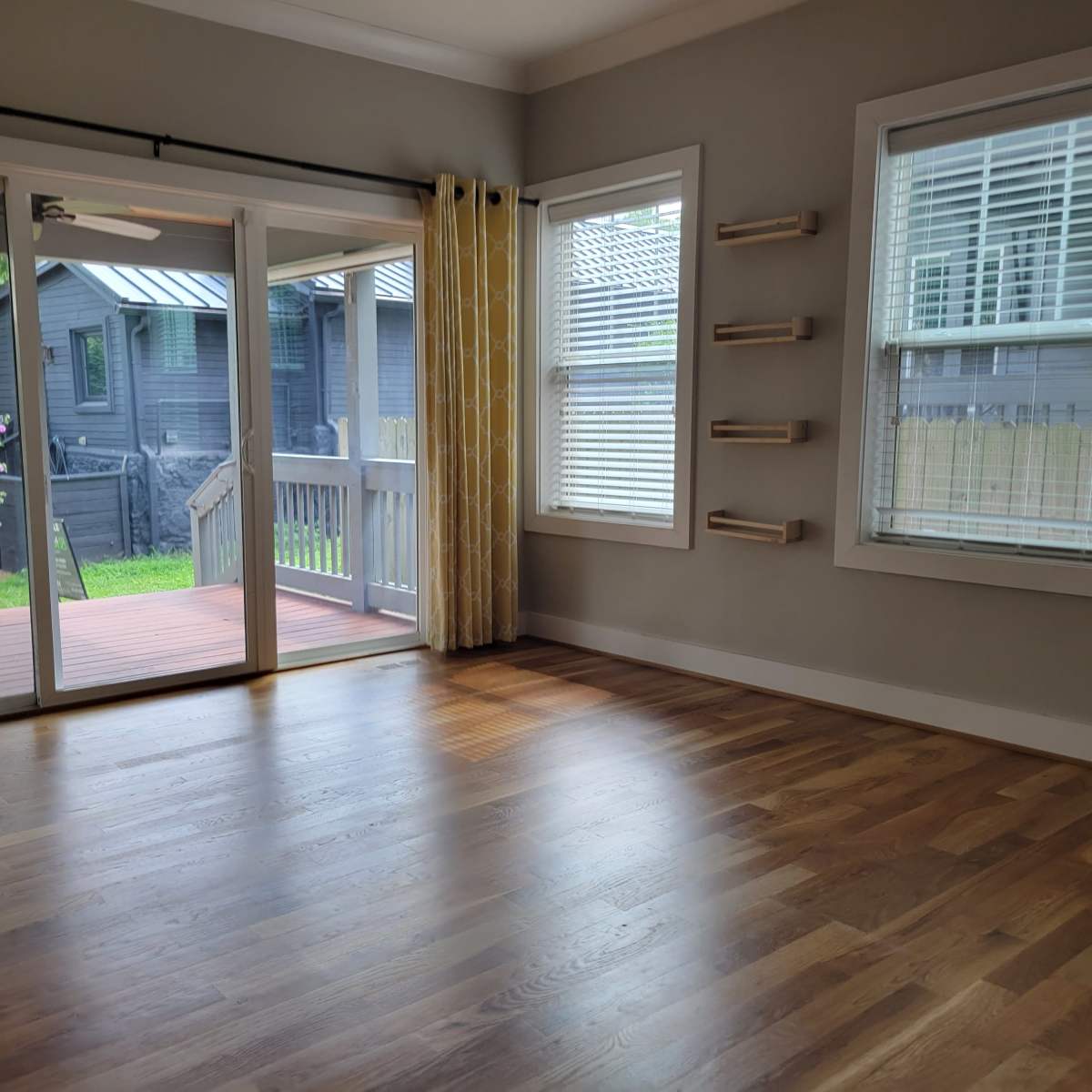 The room features hardwood floors, large windows with yellow curtains, and a sliding door that opens to a wooden deck.