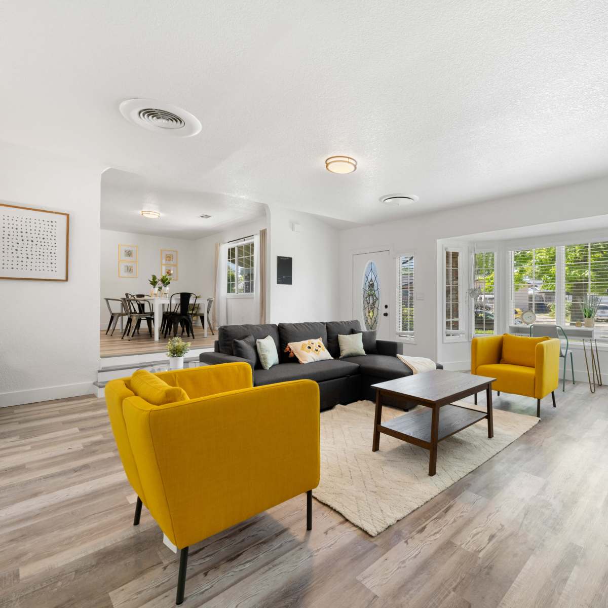 A modern living room featuring a gray sofa, two yellow armchairs, a wooden coffee table, and large windows providing ample natural light.