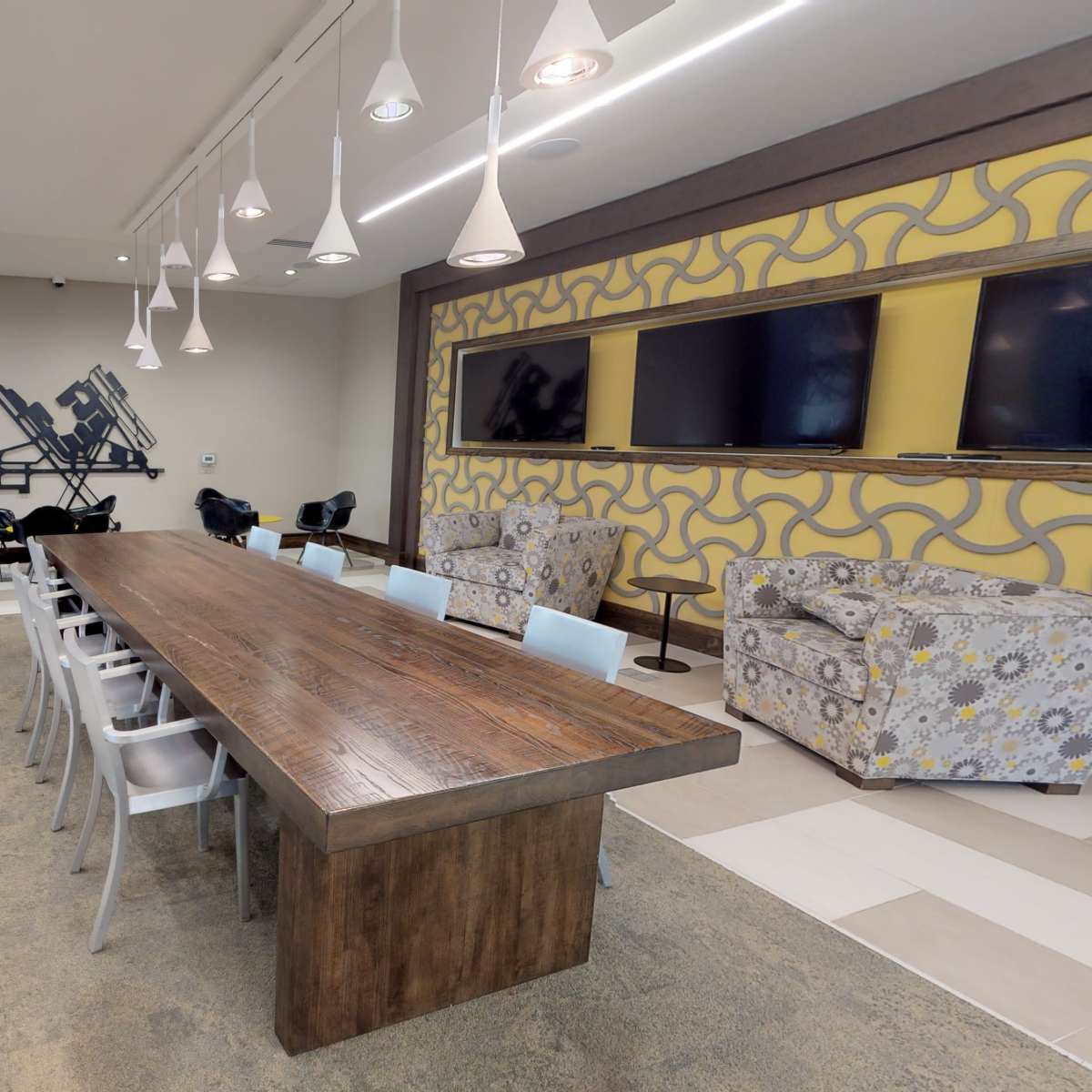 A spacious meeting room features a long wooden table, chairs, and two patterned armchairs, with a wall displaying multiple television screens.