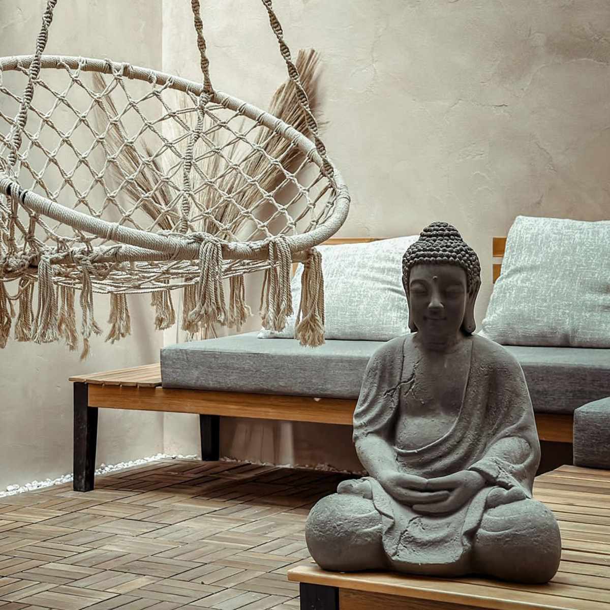 A hanging macramé chair is positioned next to a seated Buddha statue on a wooden floor in a minimalist living space.