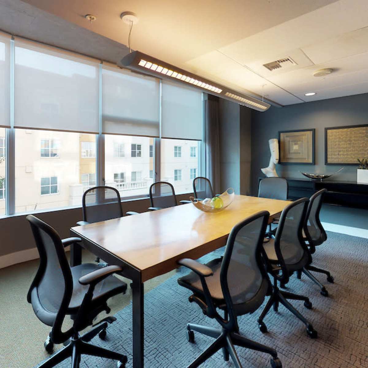 A modern conference room features a long wooden table surrounded by ergonomic chairs, with large windows providing natural light and a view of the outside.