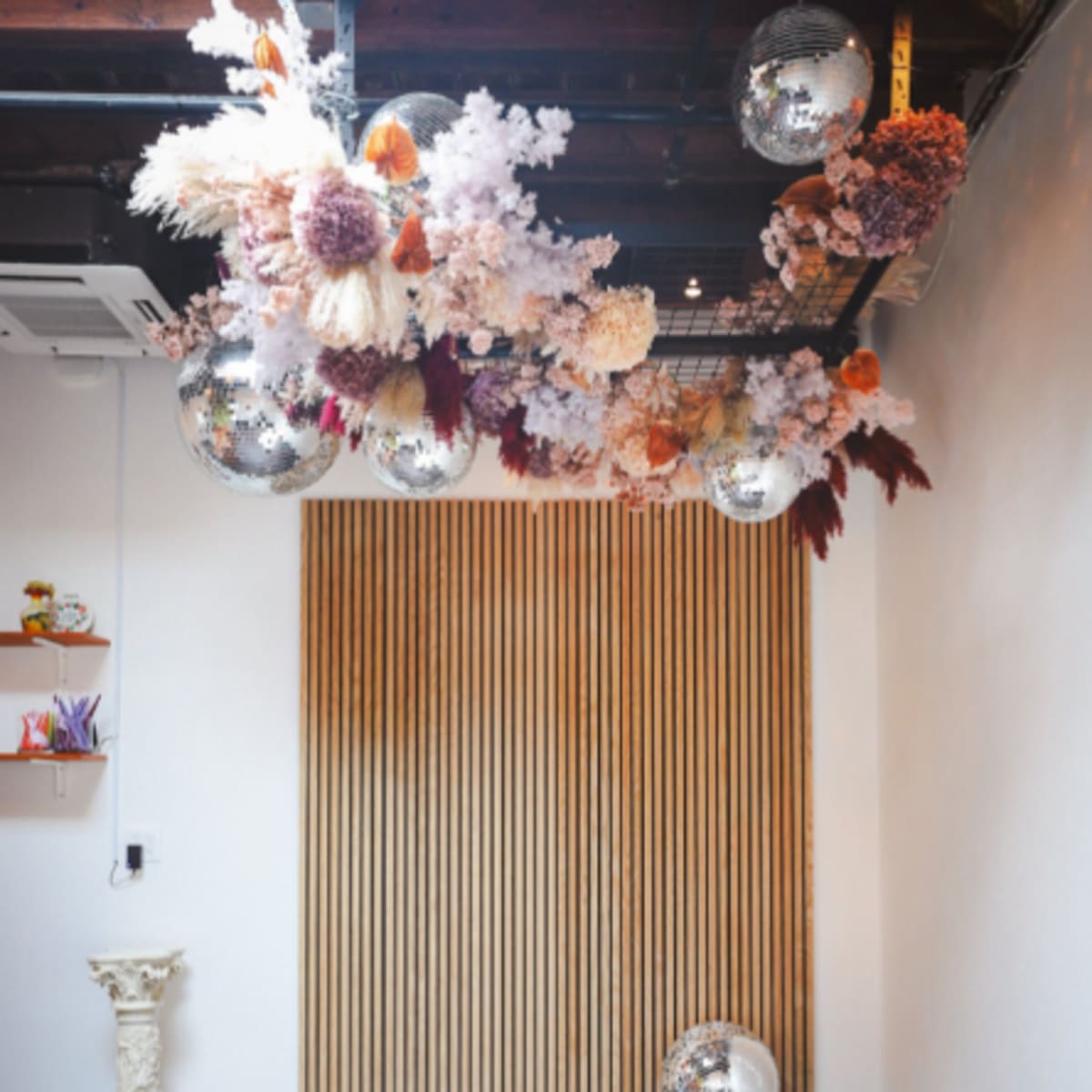 A simple interior space with white chairs arranged in front of a wooden panel backdrop and decorative floral arrangements hanging from the ceiling.