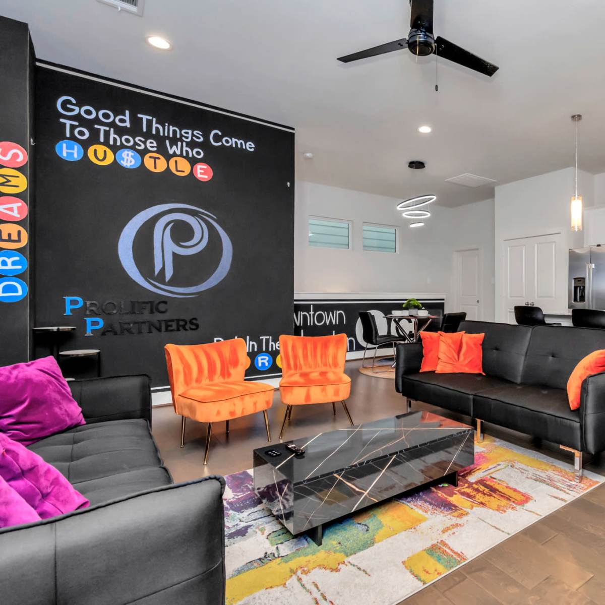 A modern living space with a black sofa, orange chairs, and a kitchen area featuring white cabinets and a colorful rug.