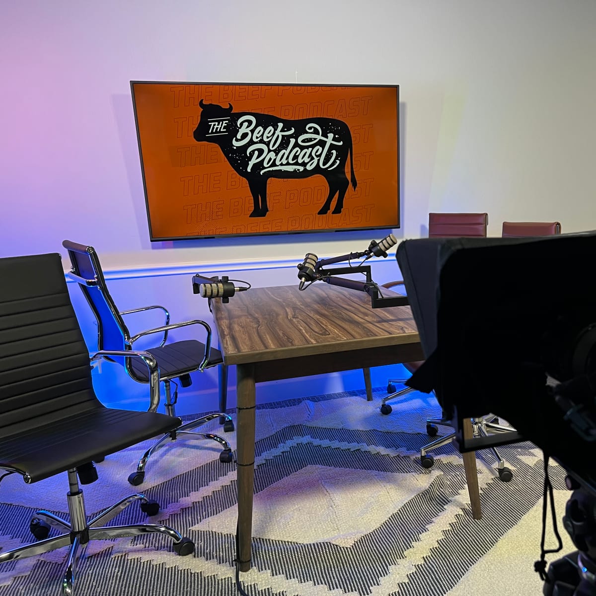 A podcast recording studio with a wooden table, two microphones, and a large screen displaying "The Beef Podcast" logo on the wall.
