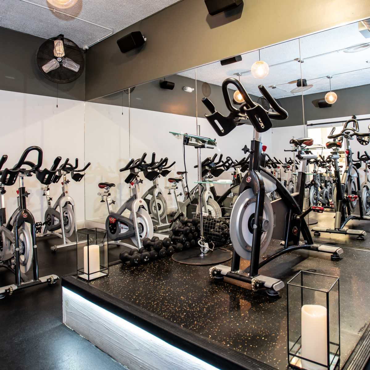 A fitness studio with multiple stationary bicycles arranged in rows, facing a mirrored wall, and equipped with some weights and ambient lighting.