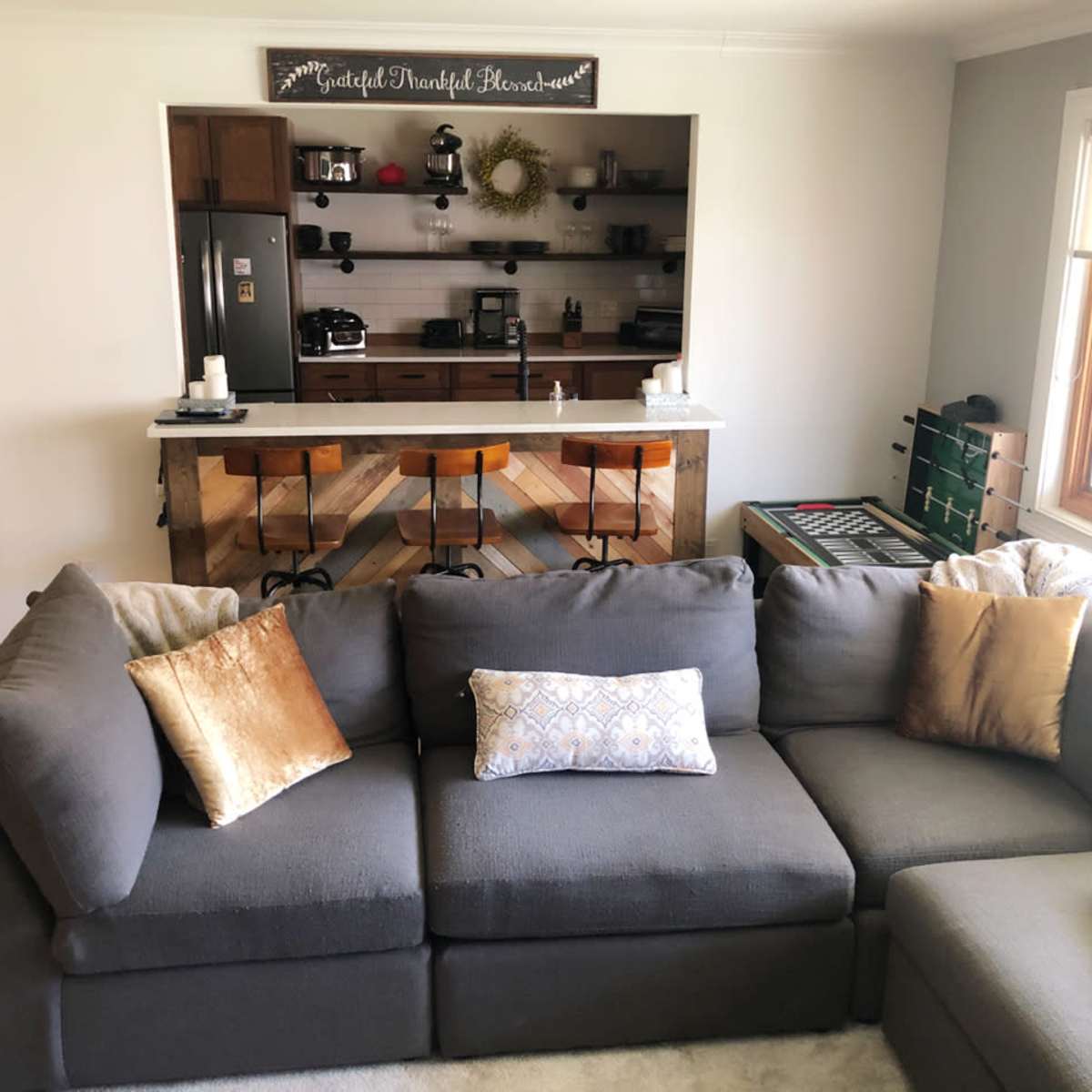 A cozy living room with a dark gray sectional sofa, a small wooden coffee table, and a view of a kitchen area with a bar and various appliances in the background.