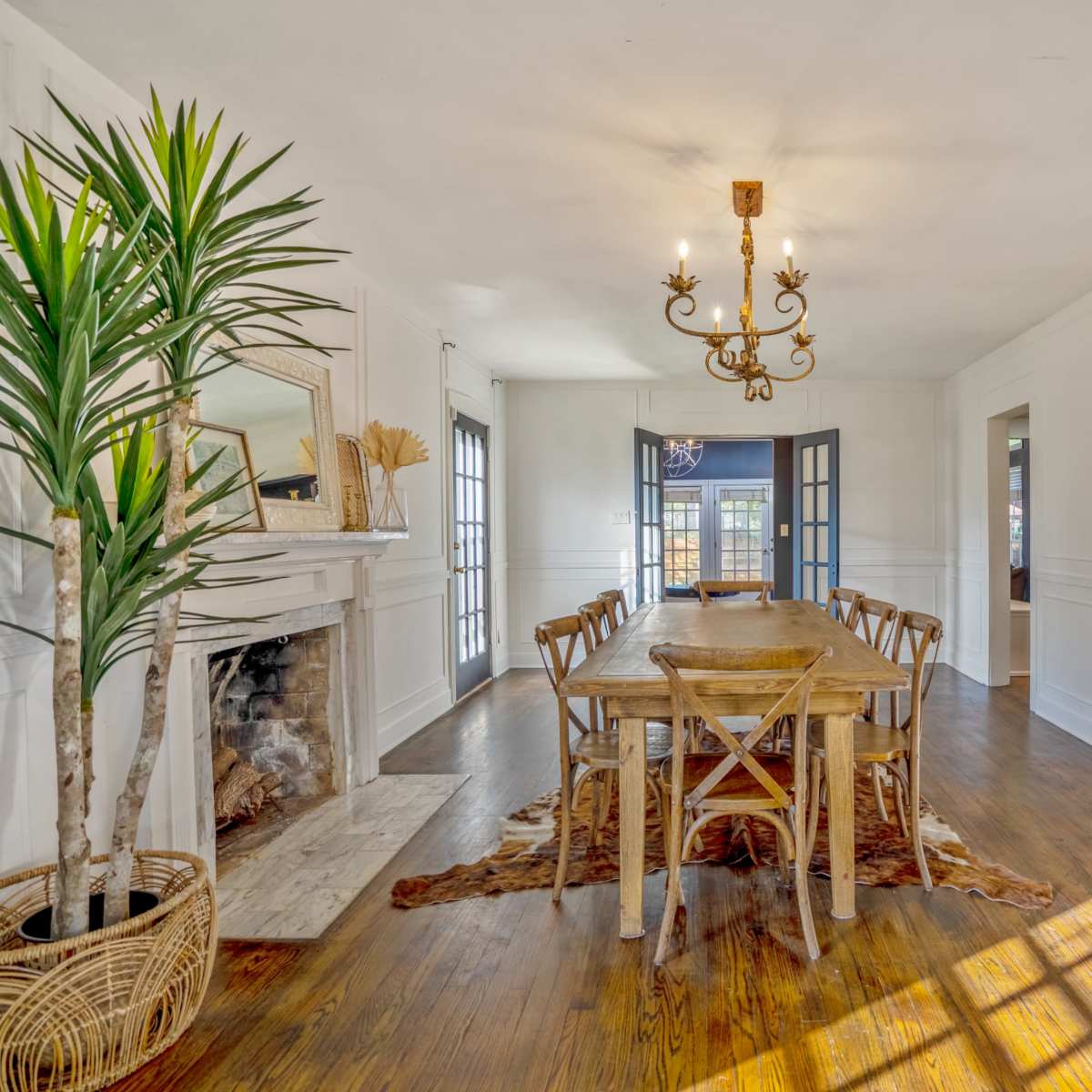 A spacious dining room features a long wooden table surrounded by chairs, a chandelier hanging above, and a decorative fireplace against a white wall.
