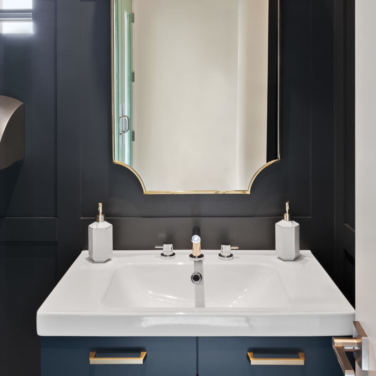 A modern bathroom vanity with a large mirror, wall-mounted light fixture, and a dual sink countertop against a dark wall.
