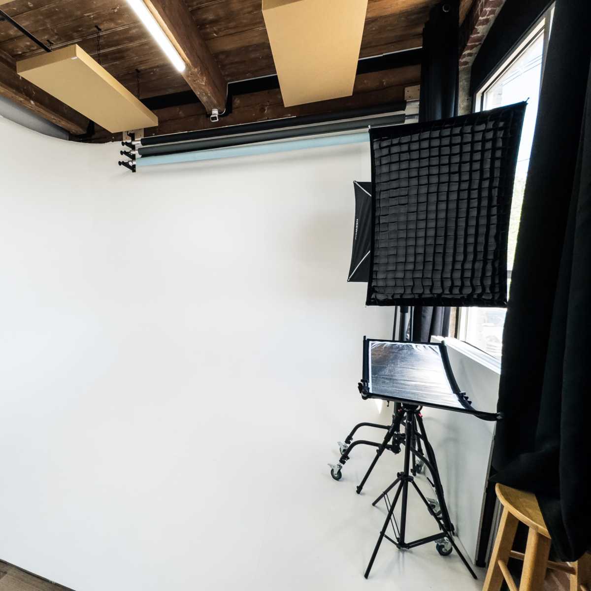 A photography studio with a white backdrop, lighting equipment on a stand, and wooden beams on the ceiling.