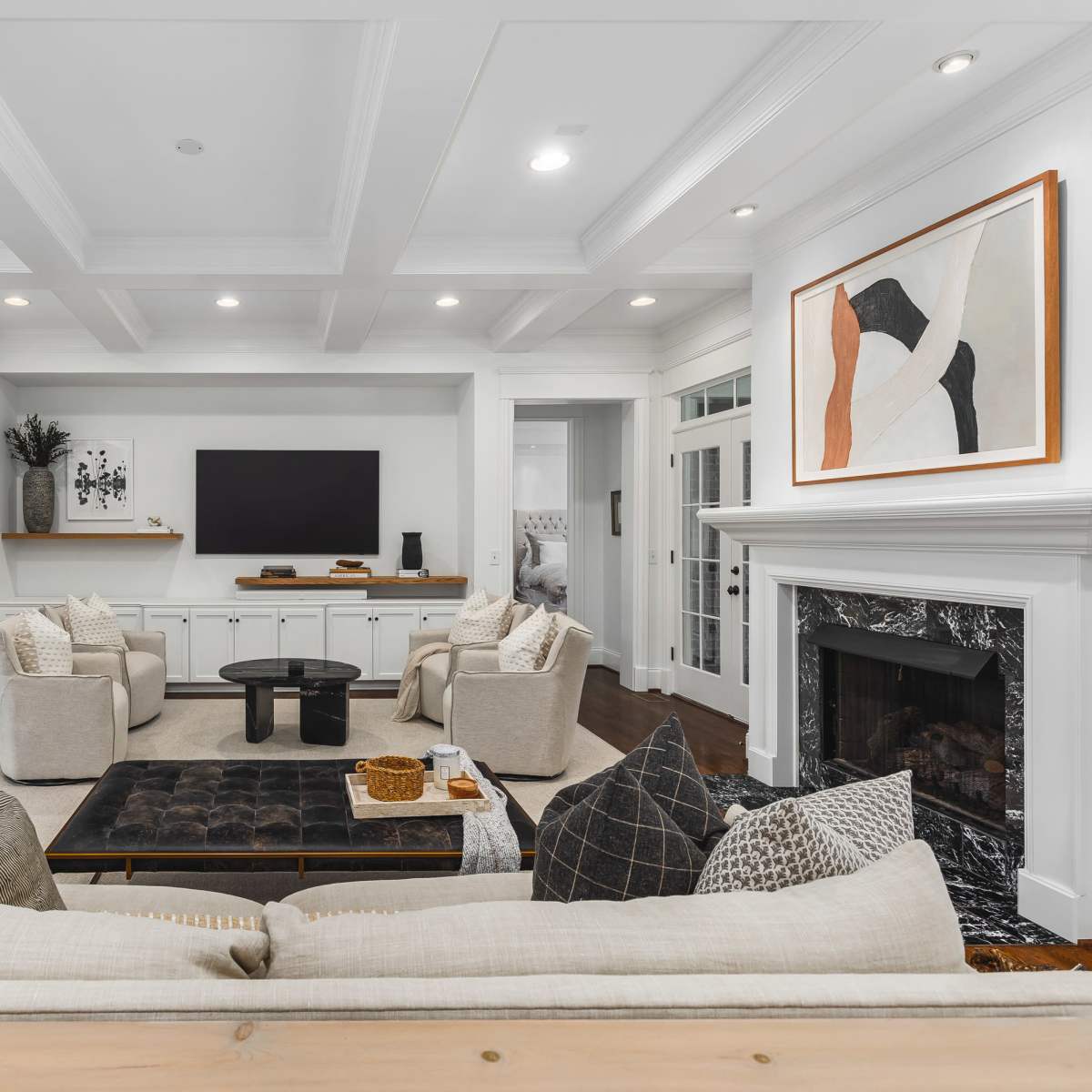 A modern living room features a central coffee table surrounded by plush armchairs and a fireplace, with a television mounted on the wall and artwork displayed nearby.