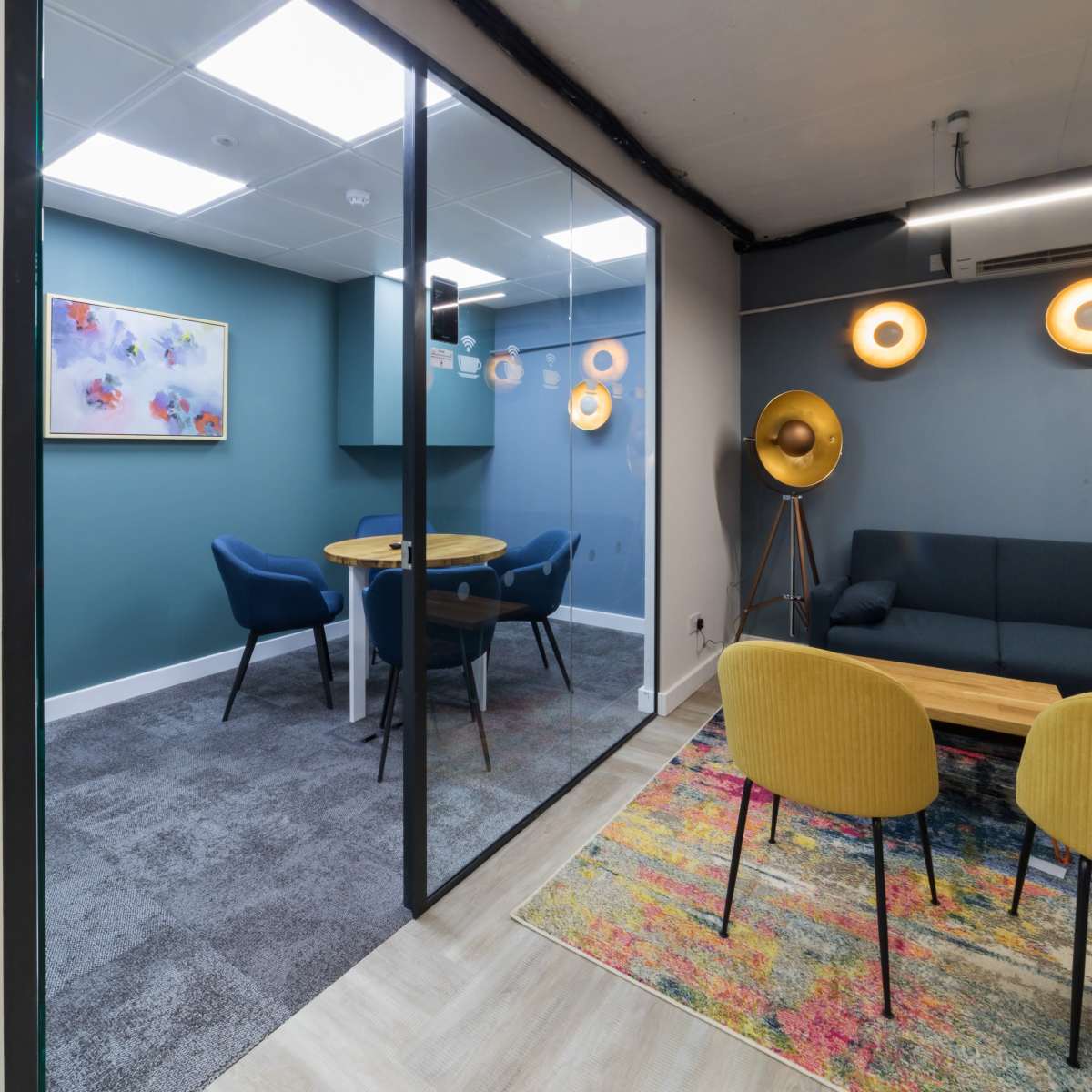 A modern office space featuring a glass-walled meeting room with a wooden table and chairs, alongside a lounge area with a sofa and decorative lighting.