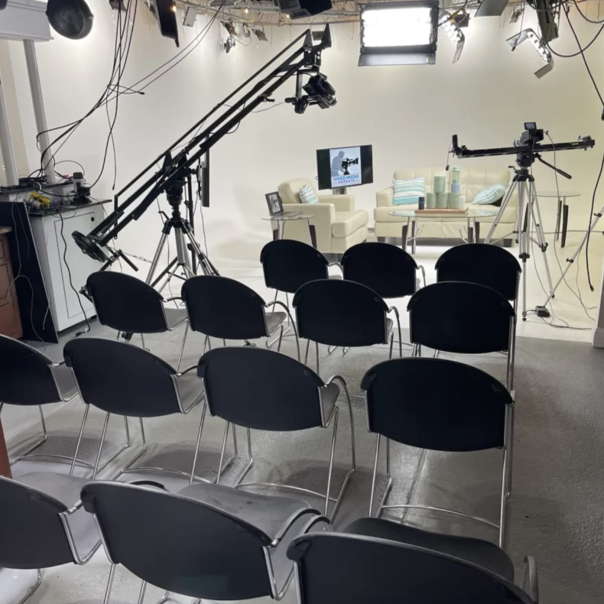 A studio setup with several empty black chairs arranged in rows facing a set featuring a couch and cameras on tripods.