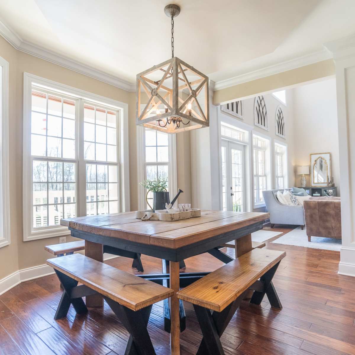 A wooden dining table with bench seating is positioned under a geometric chandelier in a bright, open dining area with large windows.