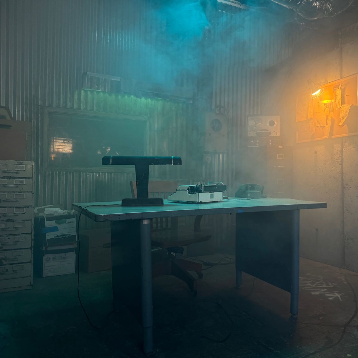A small, dimly lit office with a table and chair, surrounded by filing boxes and illuminated by a yellow light in the background.