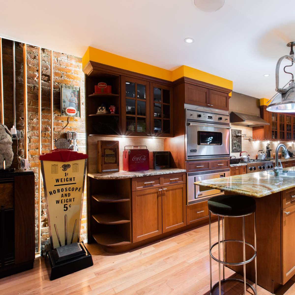 A modern kitchen featuring wooden cabinets, granite countertops, industrial-style light fixtures, and a mix of orange and brick walls.