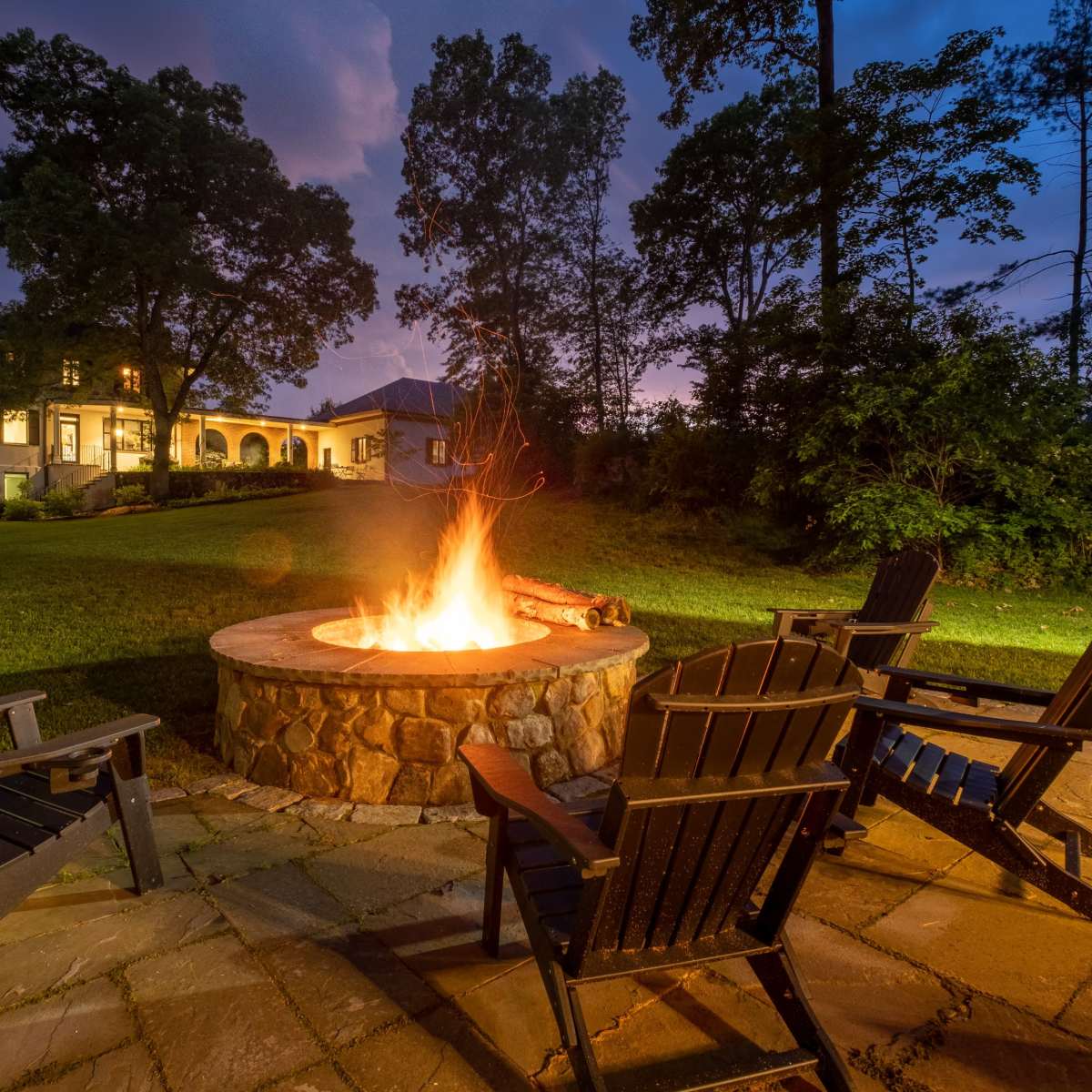 A stone fire pit surrounded by black wooden chairs is positioned on a patio in a yard with a large house and trees in the background during twilight.